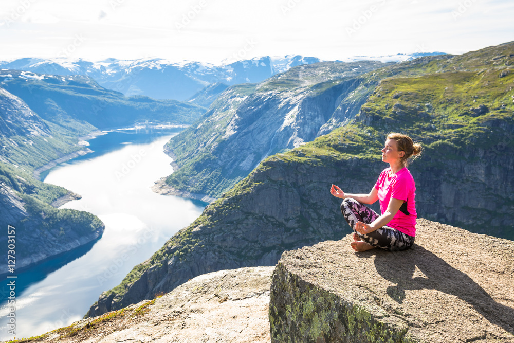 Fototapeta premium Young woman doing yoga on Trolltunga. Happy girl enjoy beautiful lake and good weather in Norway.