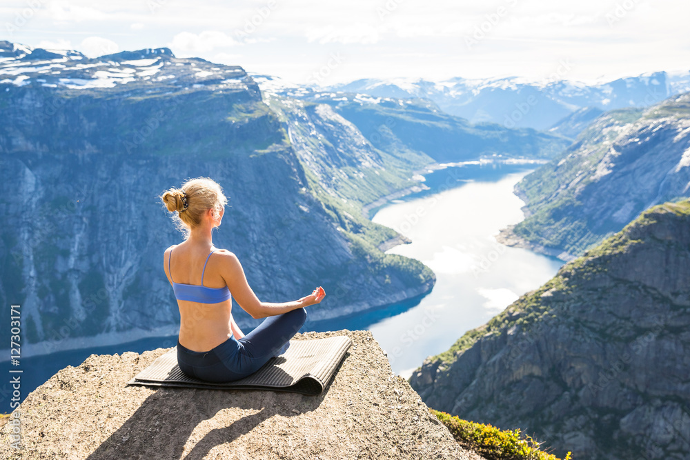 Young woman doing yoga on Trolltunga. Happy girl enjoy beautiful lake and good weather in Norway.