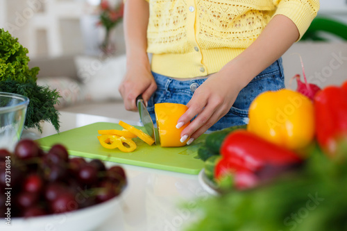 The hands of a young slender woman  clad in a yellow blouse and blue jeans  working on a large bright kitchen slicing fresh vegetables for the preparation of dietary salad a large kitchen knife