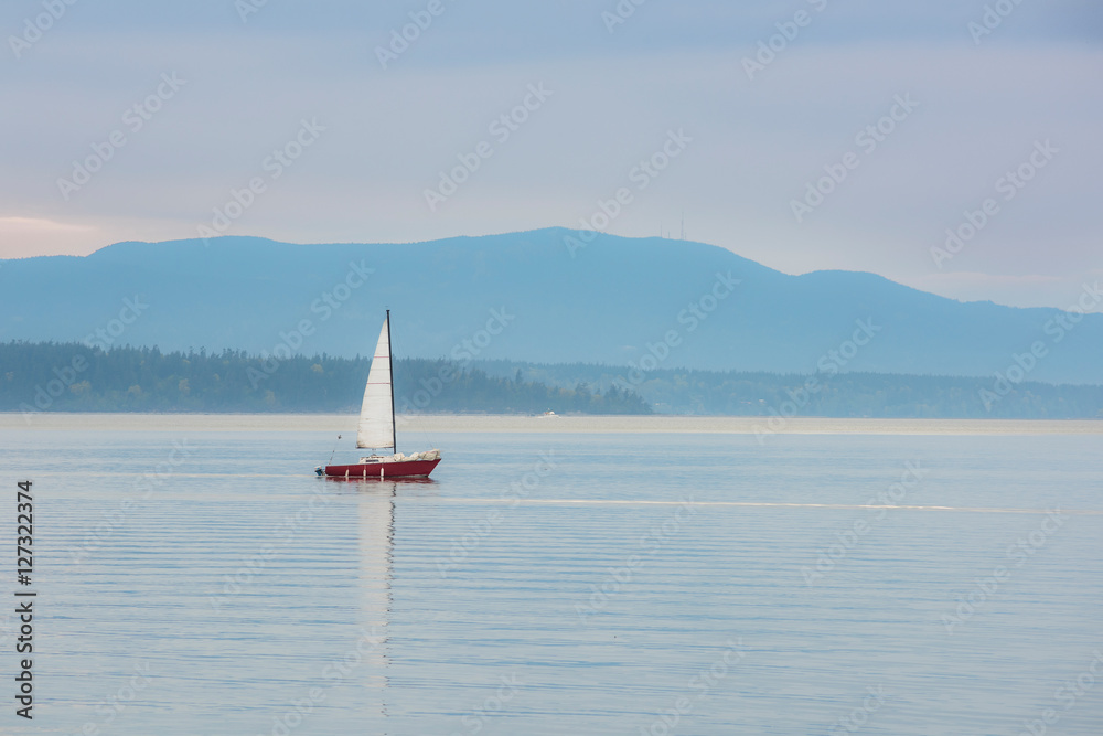 Red sailboat sailing in calm blue bay in Bellingham, Washington during purple sunset with mountain
