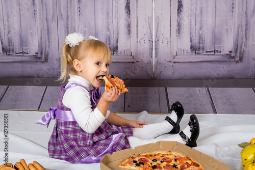 The child, a little girl sitting on the floor and having fun eating pizza. Unhealthy diets.