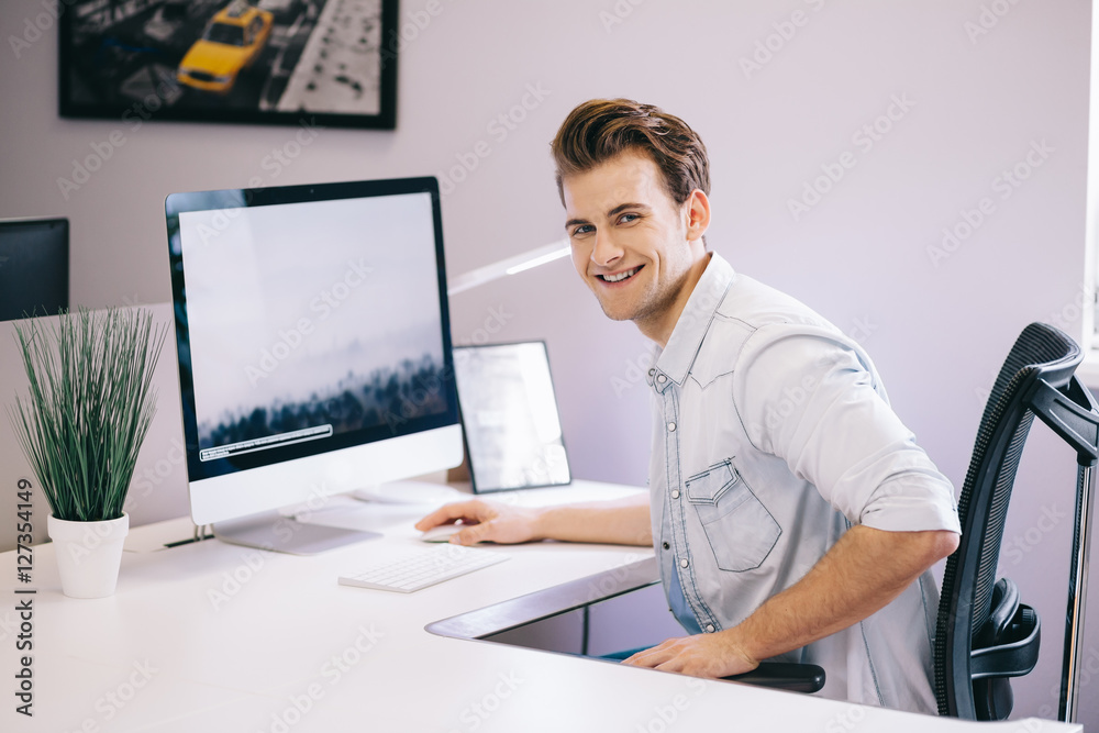 Young worker sitting in an office at the computer. Freelancer in a white shirt. The designer sits in front of window in the workplace.