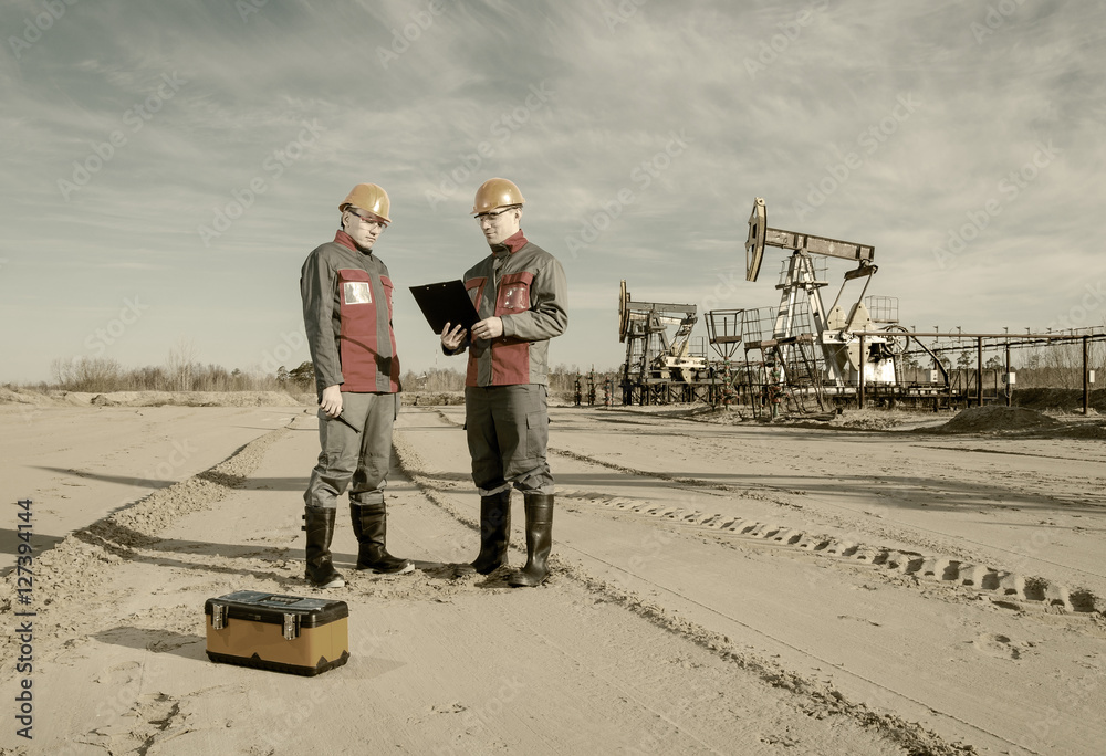 Two workers in the oilfield, one holding the radio second showing papers. Pump jack and wellhead background. Oil and gas concept. Toned. Two workers in the oilfield, one holding the radio second showing papers. Pump jack and wellhead background. Oil and gas concept. Toned.