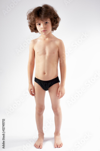 Naked curly-haired boy in shorts standing on the floor and looking into the camera. Gray background.