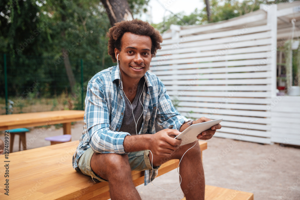 Fototapeta premium Cheerful african young man listening to music from tablet