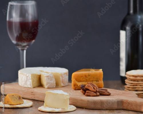 Brie, pepper jack, pecan halves and crackers on wooden cheese board, with one bottle and one glass of red wine, against dark background - celebrating alone concept