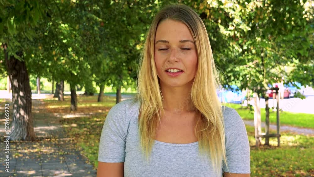 young pretty blond woman smiles to camera - park with trees in background young pretty blond woman smiles to camera - park with trees in background