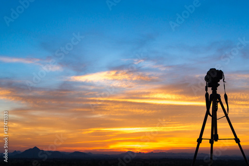 A camera set on a tripod aimed at a silhouette of a landscape at sunset