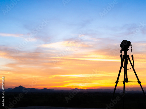A camera set on a tripod aimed at a silhouette of a landscape at sunset