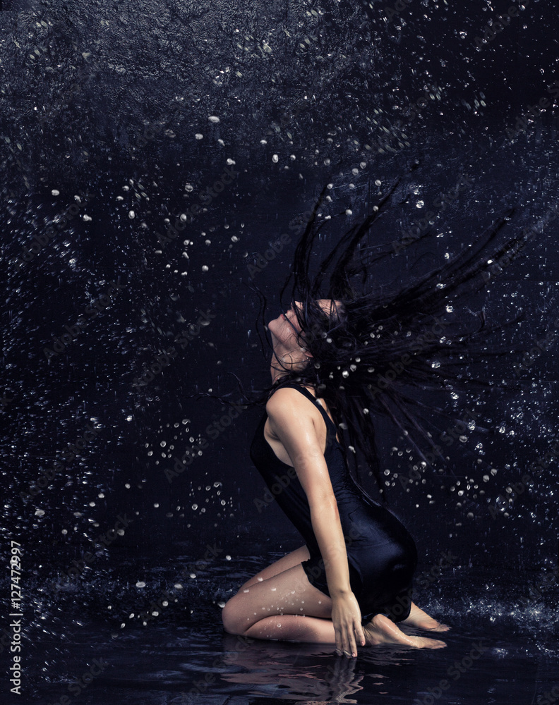 Fototapeta premium Pretty brunette woman in black dress under the water drops in aquastudio sitting in water on the knees
