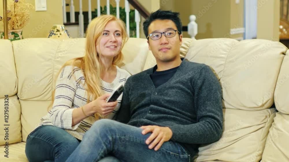 Multiethnic couple watching TV at home. Asian man and Caucasian woman. Sit on the couch, keep the remote in hand Multiethnic couple watching TV at home. Asian man and Caucasian woman. Sit on the couch, keep the remote in hand