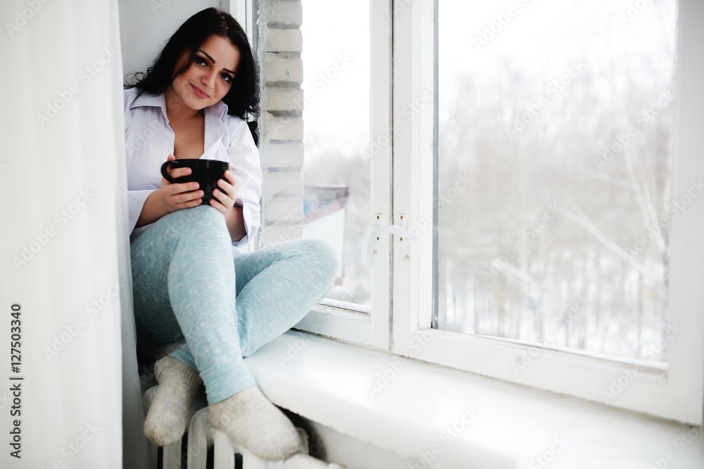 Young woman sits on window sill with cup in hands and looks out of window. Morning of winter. Young woman sits on window sill with cup in hands and looks out of window. Morning of winter.
