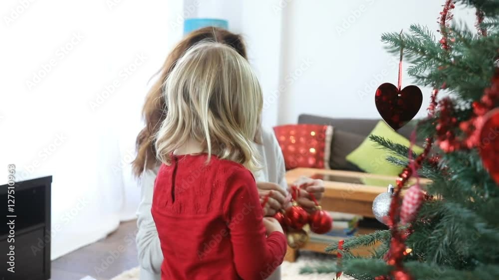 three years blonde child and brunette woman mother, at home, decorating Christmas tree, with red balls, stars and heart shapes three years blonde child and brunette woman mother, at home, decorating Christmas tree, with red balls, stars and heart shapes