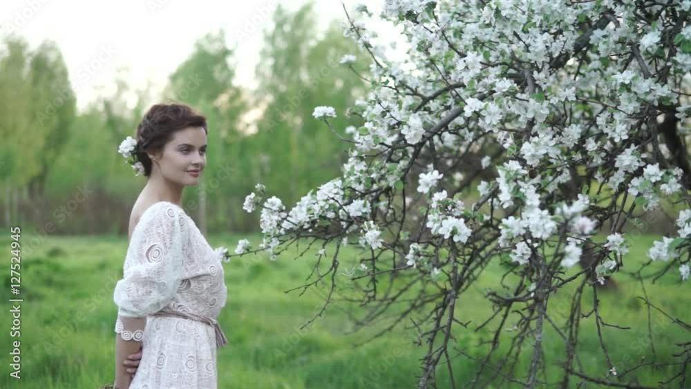 Young pretty smiling caucasian woman in white evening dress and blossom in her hair stands in a blooming spring garden. Youth, freshness, fashion, beauty, happiness concept. Young pretty smiling caucasian woman in white evening dress and blossom in her hair stands in a blooming spring garden. Youth, freshness, fashion, beauty, happiness concept.