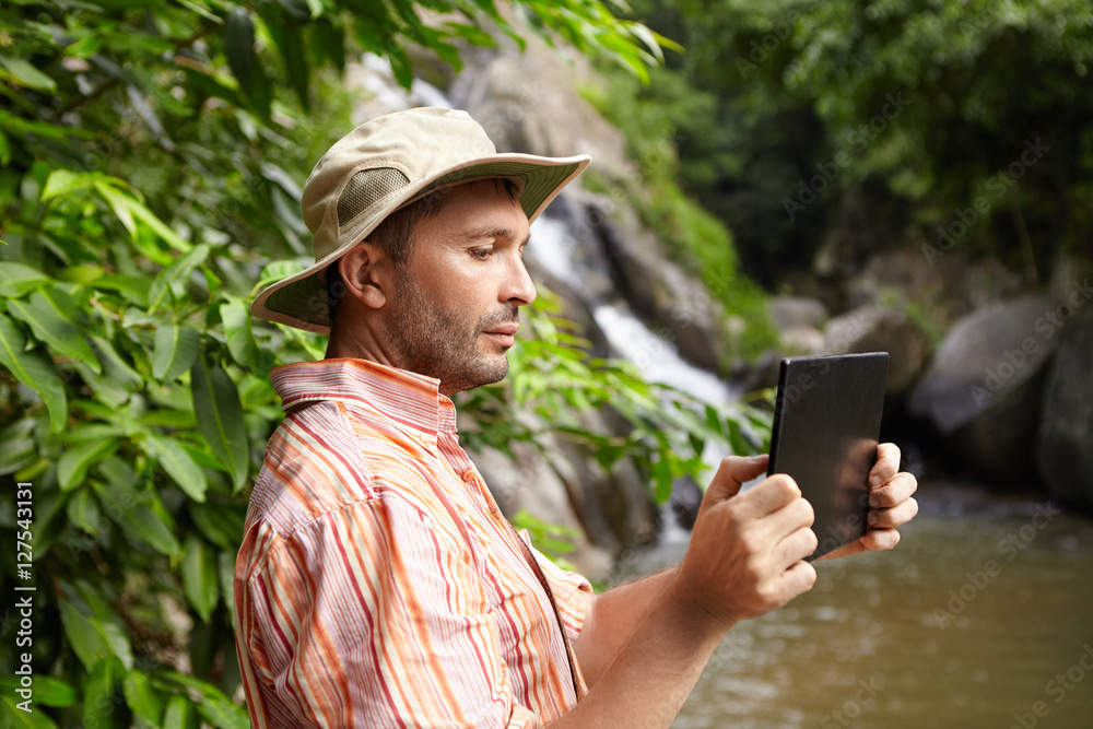 People and technology concept. Side view of bearded ecologist in panama hat taking photographs or shooting video of wild nature in tropical forest using digital tablet, standing at mountain river People and technology concept. Side view of bearded ecologist in panama hat taking photographs or shooting video of wild nature in tropical forest using digital tablet, standing at mountain river