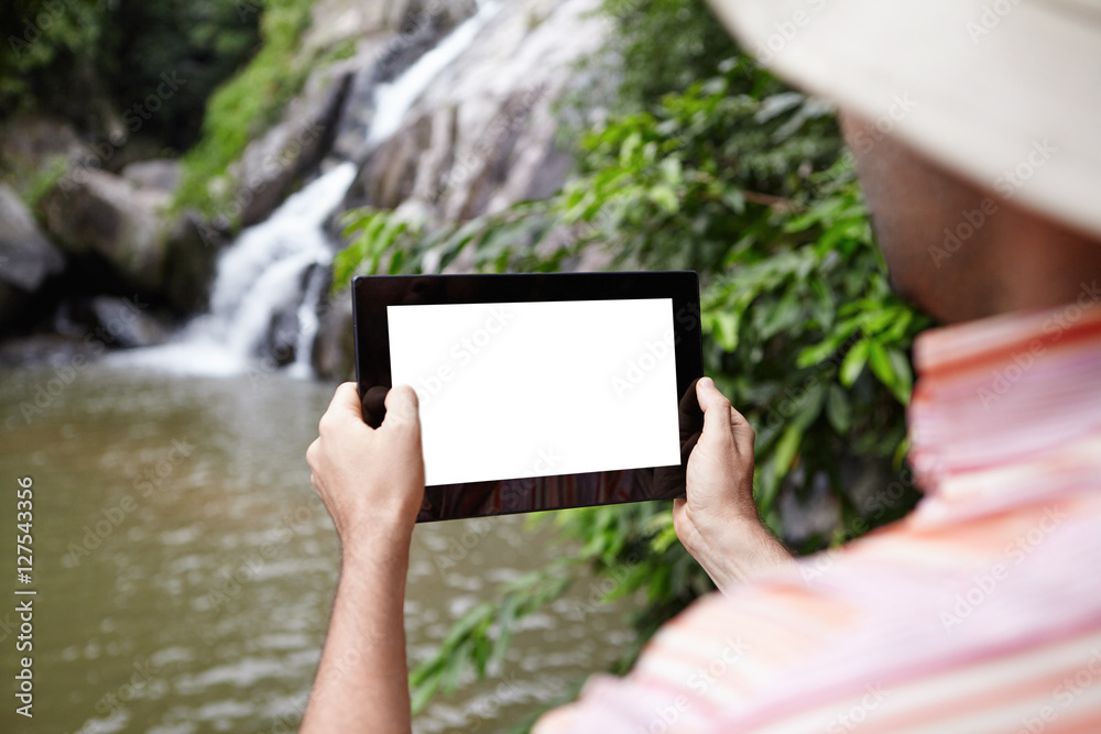 Rear view of biologist wearing panama hat taking picture of mountain waterfall in nature park using touch pad. Selective focus on blank screen with copy space for your text or promotional content