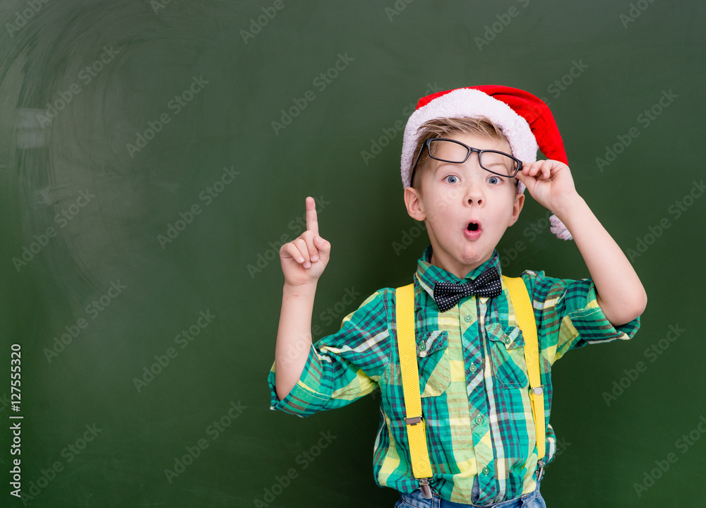 surprised boy in red christmas hat near a green chalkboard showing finger up