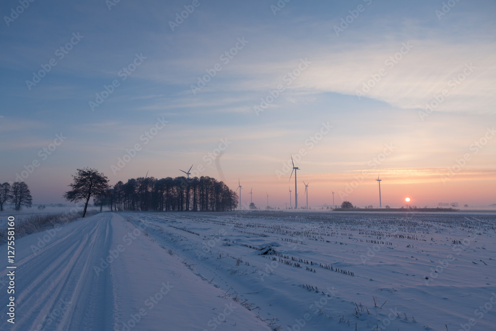 Road covered with snow is leading to wind turbines with sunset