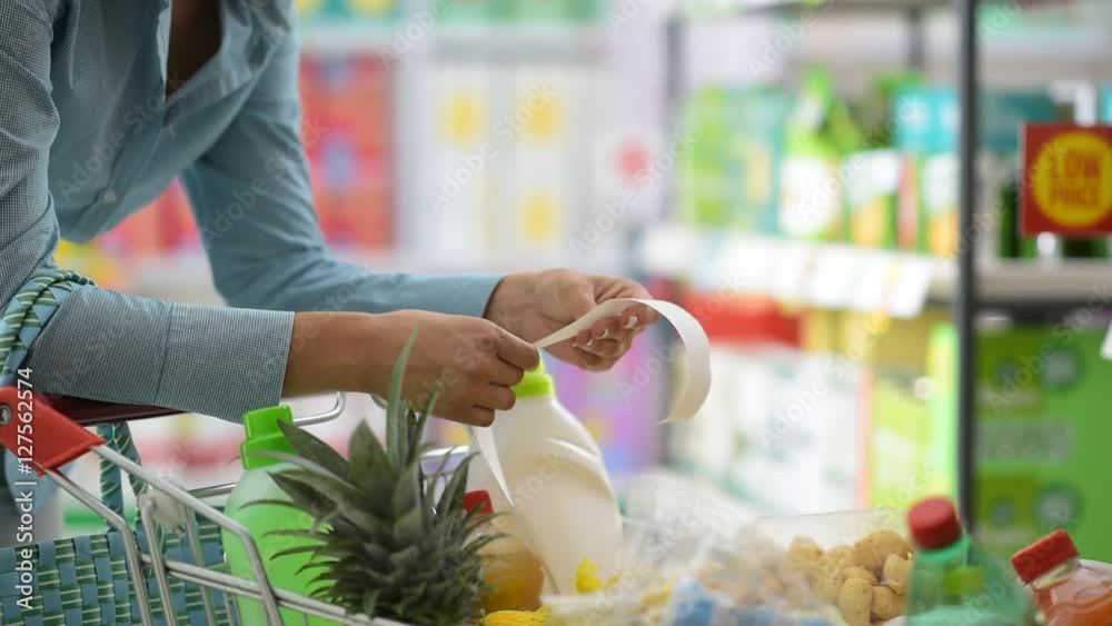 Woman doing grocery shopping at the supermarket, she is leaning on a full shopping cart and checking a long receipt