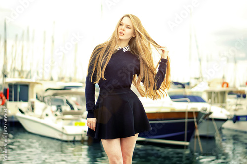 Beautiful blonde cute caucasian woman posing in harbor with luxury yachts on background. Girl wearing black fashionable skirt, holding her hair.