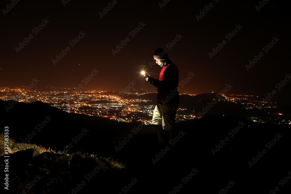 Young man with phone on top of the hill observing the night city view.