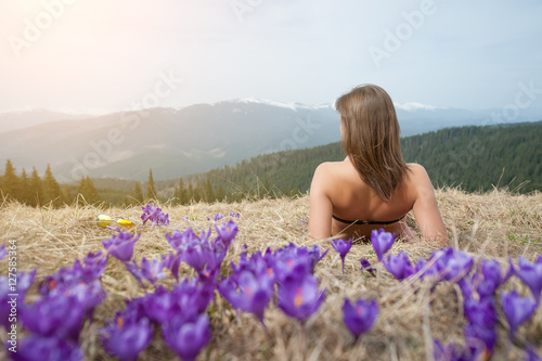 Back view of naked woman in swimsuit is lying on the spring grass nearly crocuses in the mountain. Beautiful landscape, snowy mountains on the background