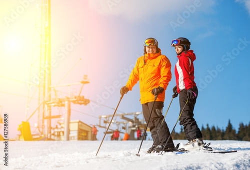 Couple holding skis standing together at a winter resort with ski lifts and blue sky in background. Man is wearing orange jacket, female in red jacket, both is wearing helmet and goggles.