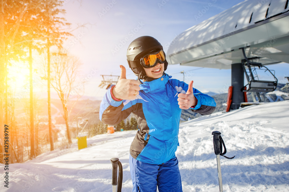 Portrait of young beautiful female skier at ski resort smiling and showing thumbs up. Winter sports concept. Woman is wearing blue jacket and blue pants, helmet and orange goggles. Bukovel, Ukraine - obrazy, fototapety, plakaty 