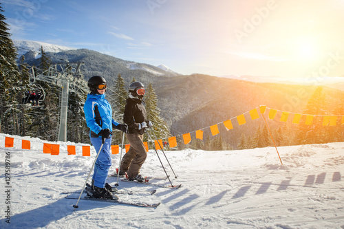 Young couple of women enjoying skiing at ski resort on a sunny day against ski lift and mountains background. Carpathian Mountains, Bukovel, Ukraine