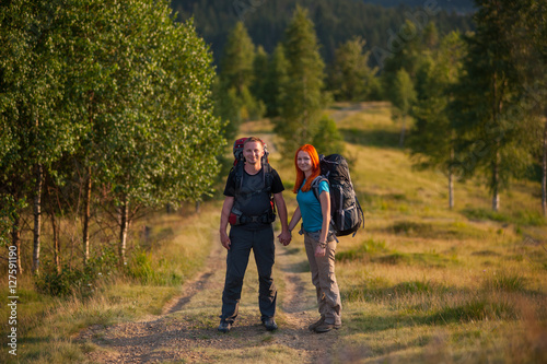 Young tourists with backpacks holding hands on a trail to the mountain against trees and forest in the distance on the background © anatoliy_gleb