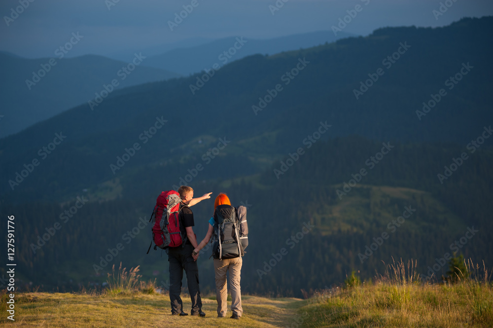 Rear view of happy couple tourists with backpacks holding hands and standing along a road with beautiful mountain landscape on background. Man is showing mountains