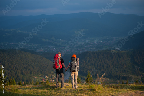 Rear view pair with backpacks holding hands standing on the ridge of hill, enjoying the view of beautiful open overlook on the mountains and village in the valley
