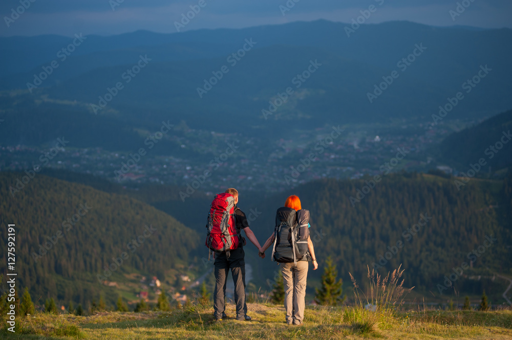 Rear view couple hikers with backpacks holding hands, standing on the top of hill, enjoying the view of beautiful open overlook on the mountains and village in the valley