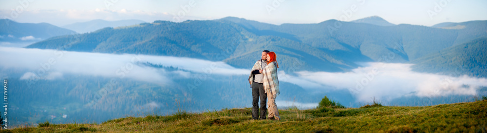 Sweety couple hikers covered with a blanket standing together on a hill, enjoying beautiful mountain landscape with morning haze over the mountains and forests. Panorama