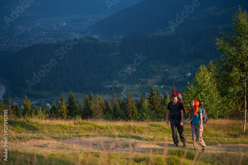 Couple of hikers with backpacks walking in the beautiful mountains area, holding hands and looking to each other. Lifestyle active vacations concept mountains landscape on background