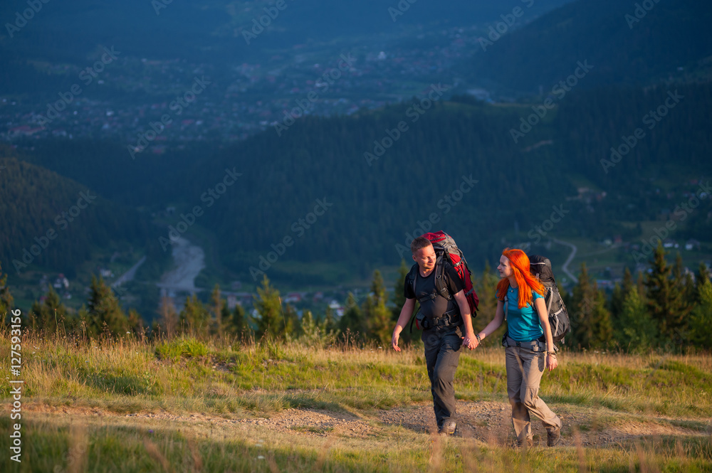 Happy couple of tourists with backpacks hiking in the beautiful mountains area, holding hands and looking to each other. Lifestyle active vacations concept mountains landscape on background