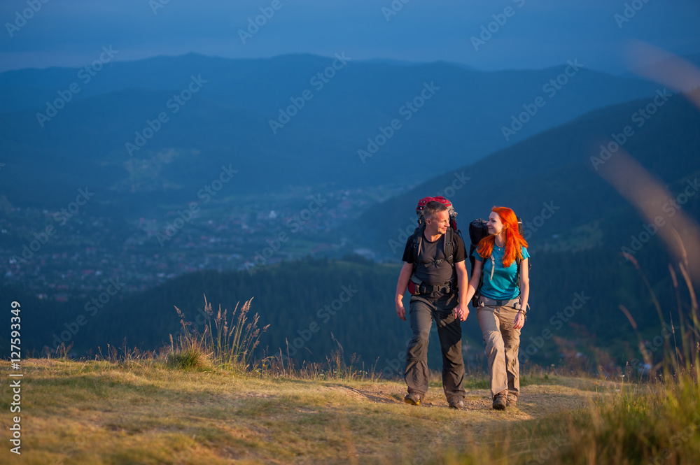 Tourists man and woman with backpacks walking in the beautiful mountains area, holding hands and looking to each other. Lifestyle active vacations concept mountains landscape on background Tourists man and woman with backpacks walking in the beautiful mountains area, holding hands and looking to each other. Lifestyle active vacations concept mountains landscape on background