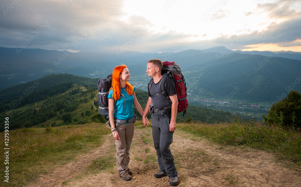 Happy man and red-haired woman standing, looking to each other, holding hands, smiling on the road in the mountains on the background of the landscaped mighty mountains, forests, hills and clouds sky