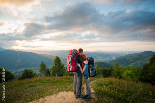 Back view of couple tourists with backpacks standing embracing and enjoying the view of beautiful open overlook on the mountains, forests, hills, village in the valley and cloudy sky