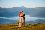 Young pair hikers standing on a hill, embracing each other against beautiful mountain landscape with morning haze over the mountains on background. Red-haired female covered with a plaid