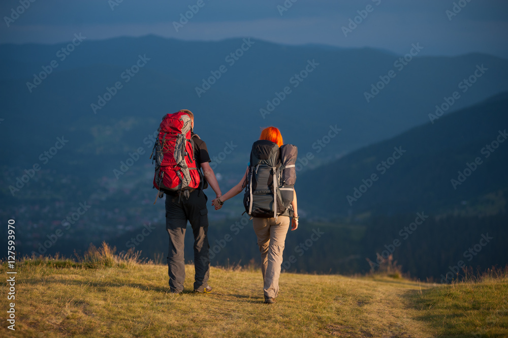 Rear view of couple hikers with backpacks holding hands and walking along a road with beautiful mountain landscape on background