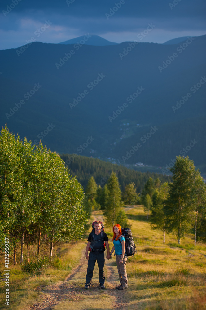 Hikers with backpacks holding hands on a trail to the mountain against trees and forest in the distance on the background. Lifestyle active vacations concept