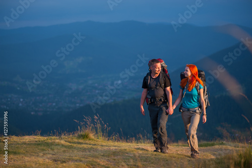 Happy couple of backpackers with backpacks hiking in the beautiful mountains area, holding hands and looking to each other. Lifestyle active vacations concept mountains landscape on background © anatoliy_gleb