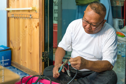 old senior asian man wear glasses, He’s sitting cross-legged and cutting wire by hand via pliers. (DIY maintenance)