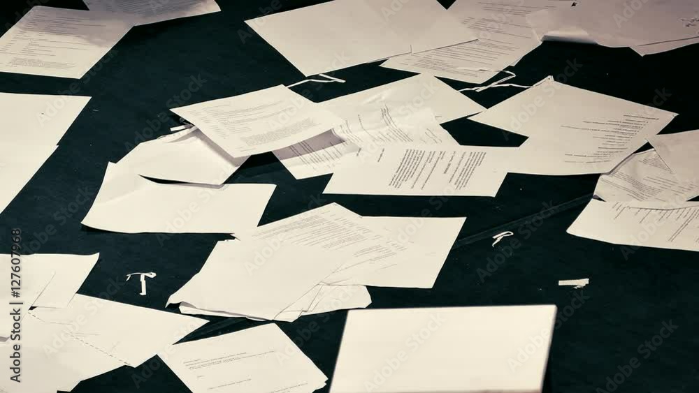 Sheets of paper with unreadable printed text fall to the black floor in a mess. People walk on the debris.