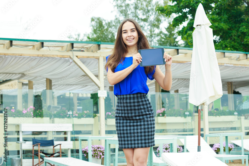 Beautiful girl with long brown hair holding a tablet computer. She is dressed in a blue blouse and plaid skirt. Girl holding tablet with two hands and smiles. The tablet screen looking at the camera.