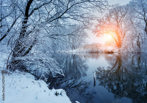 Beautiful winter in forest on the river at sunset. Winter landscape. Snowy branches on trees, beautiful river with reflection in water, sun and blue sky. Seasonal background. Frosty cold evening
