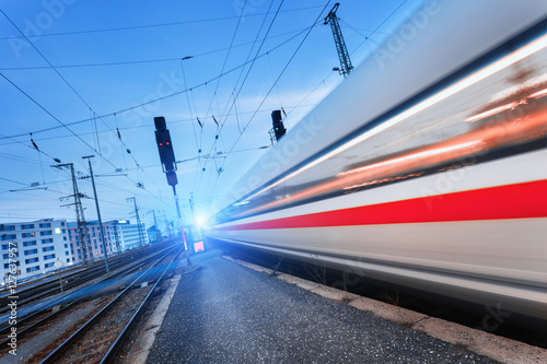 Modern high speed passenger train on railroad in motion at sunset. Blurred commuter train. Railway station at dusk with vintage toning. Travel background, railway tourism. Industrial landscape. Train