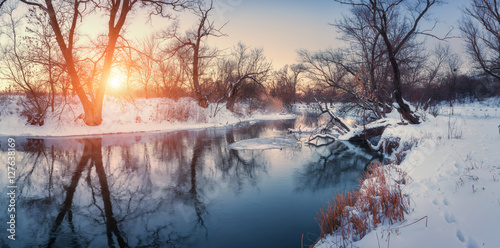 Panoramic winter landscape with trees, beautiful river at sunset. Winter forest. Season. Scenery with winter trees, water and blue sky. Christmas background with snowy forest. Reflection in water