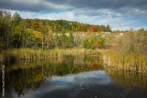 Autumn parkland featuring a sun-lit pond, vivid fall colors on trees and bright blue sky.
